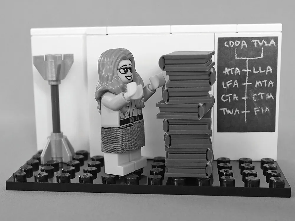 Una minifigura de LEGO de Margaret Hamilton, en blanco y negro, junto a una torre de libros de LEGO y una pizarra, recreando la foto de 1969