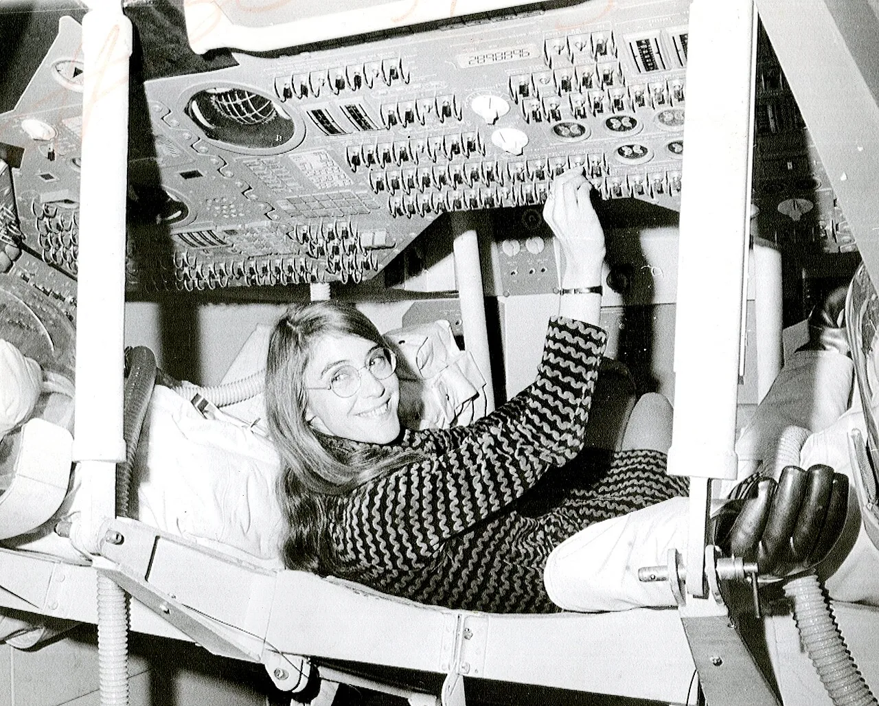 Margaret Hamilton, sonriente, tumbada en el asiento del simulador del módulo de mando del Apollo, con la mano en el panel de interruptores